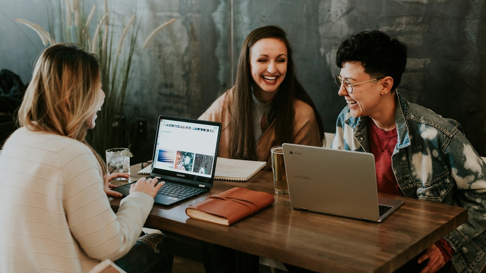 Three colleagues at a wooden table with laptops, talking and laughing, heads fully visible